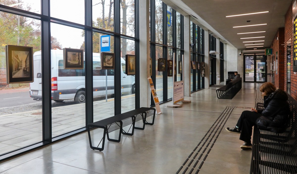Obrazy wiszące w szybach budynku centrum przesiadkowego. Na ławkach siedzą podróżni.