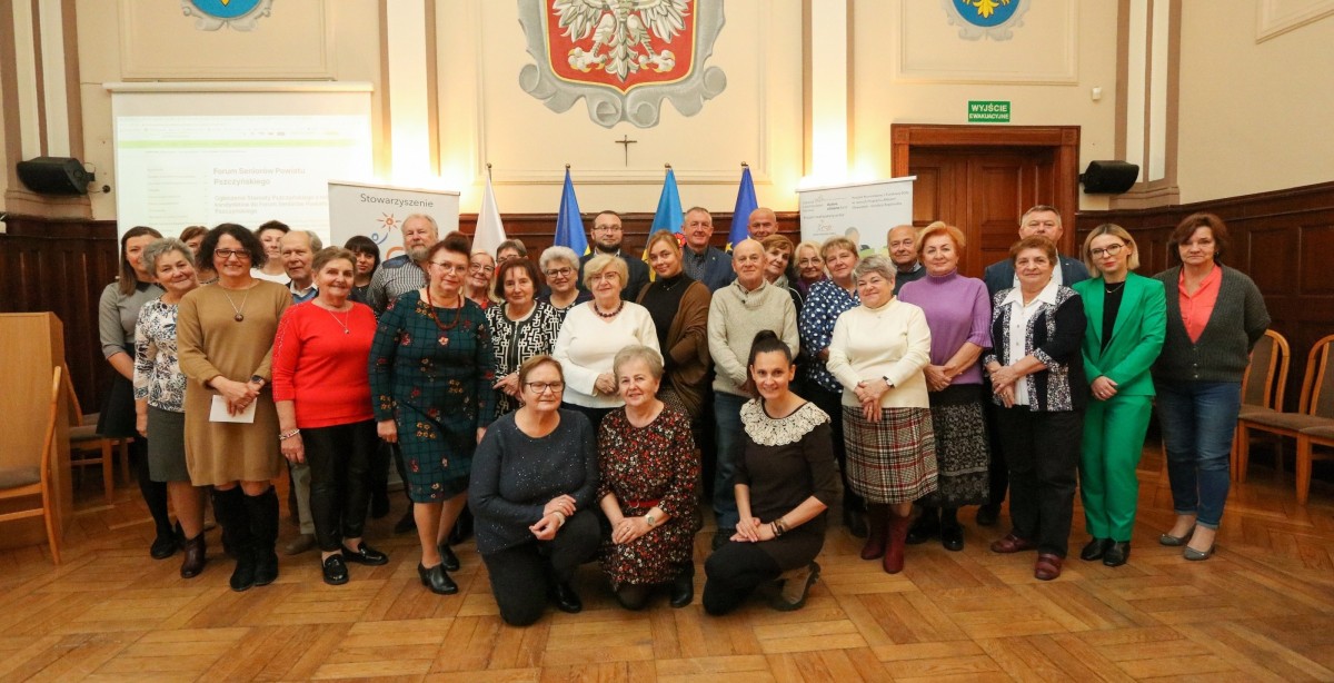 Grupa seniorów i organizatorzy spotkania w sali sesyjnej starostwa powiatowego.