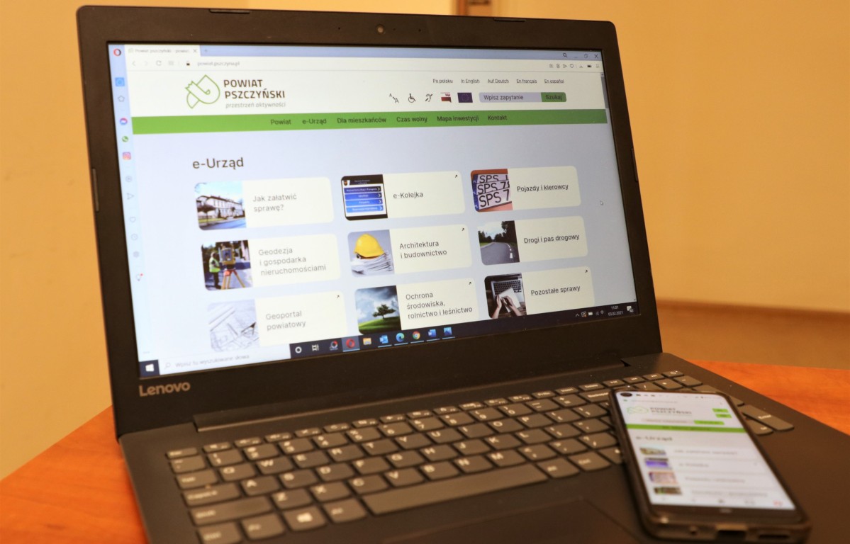 Laptop na brązowym biurku, a na nim położony telefon. Otwarta strona internetowa powiatu pszczyńskiego.