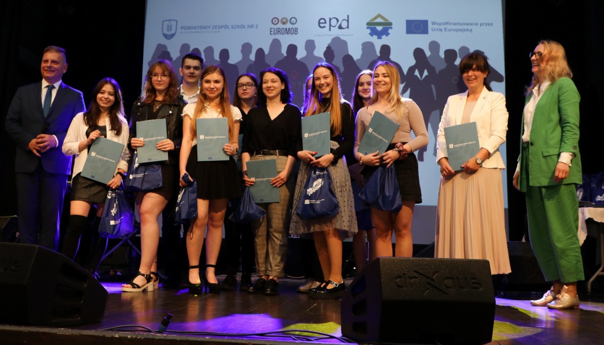 Grupa uczniów wraz z dyrektor PUP i dyrektorem stojąca na scenie z certyfikatami.