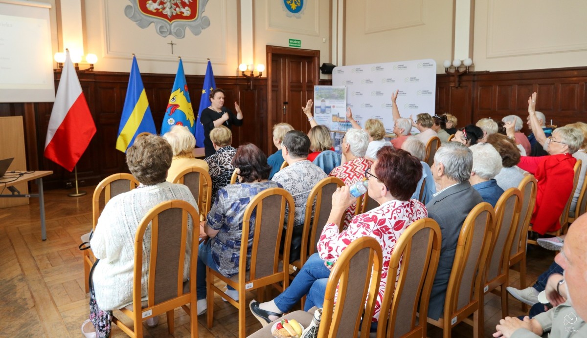 Uczestnicy konferencji siedzą na krzesłach w sali sesyjnej, przed nimi stoi prowadząca wykład.