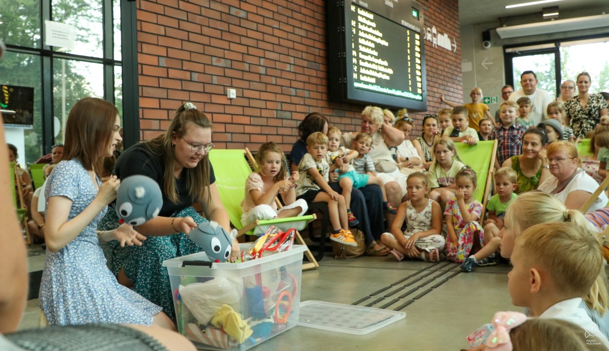 We wnętrzu centrum przesiadkowego dzieci i rodzice siedzą na leżakach i oglądają spektakl