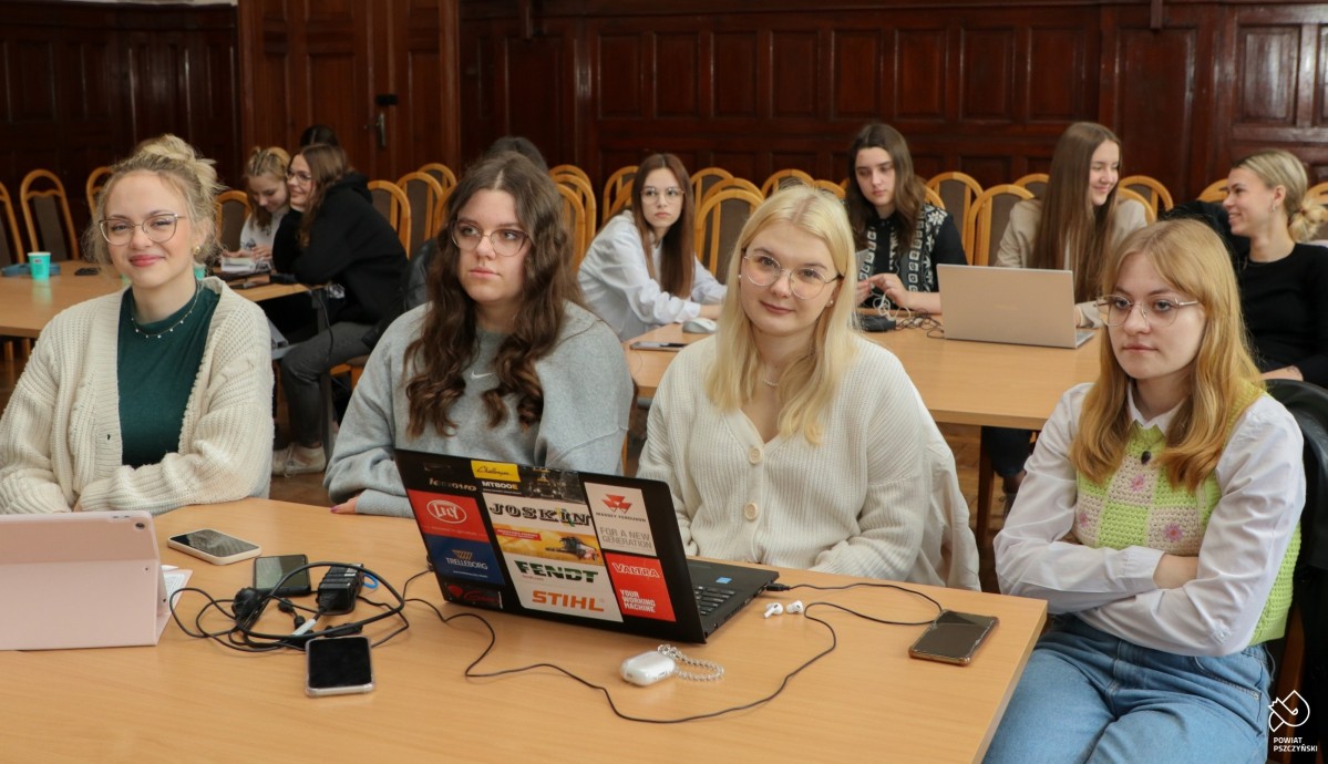 Uczennice w sali sesyjnej siedzą przy kwadratowym stole, na którym podstawiony jest laptop.
