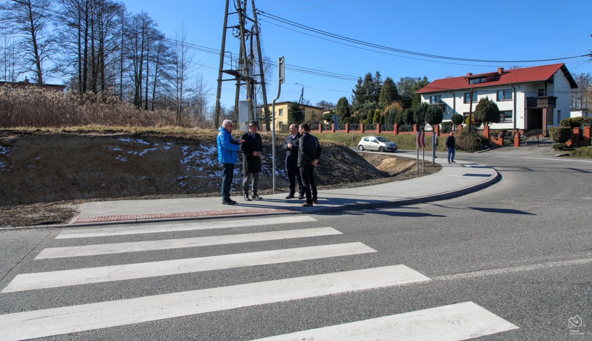 Na chodniku przy przejściu dla pieszych stoi grupa mężczyzn, samorządowców