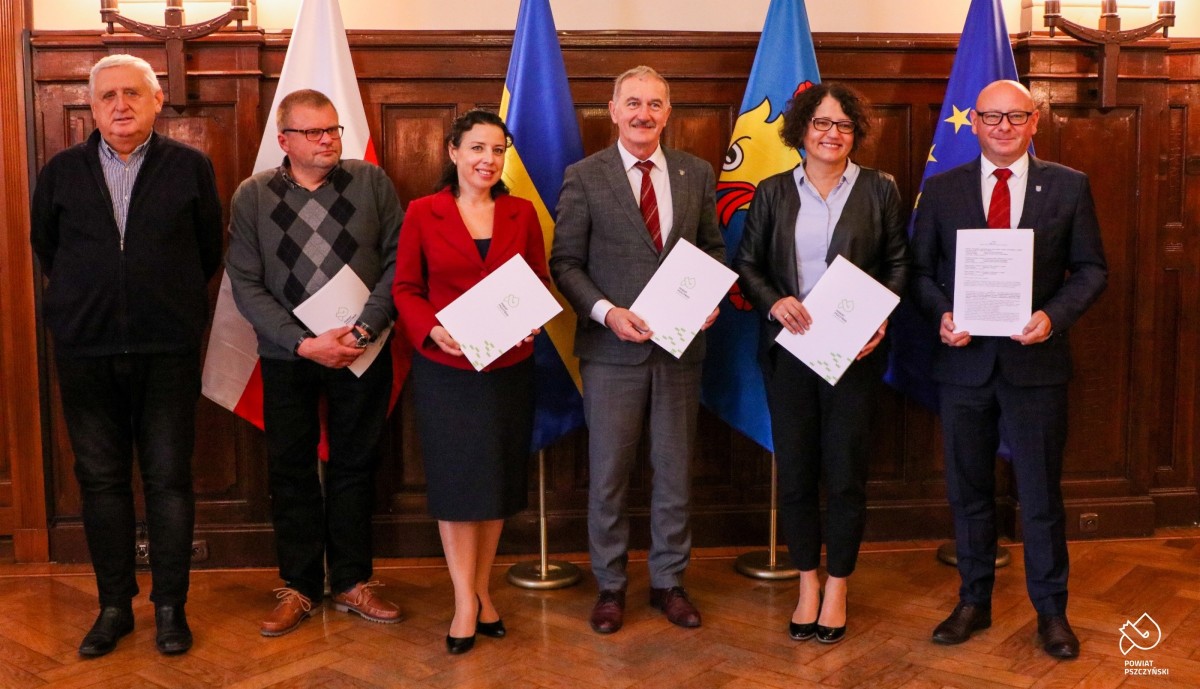 W sali sesyjnej starosta na tle flag polskiej, unijnej, wojewódzkiej oraz powiatowej stoją samorządowcy - którzy podpisali umowę w sprawie finansowania ciągu pieszo-rowerowego.