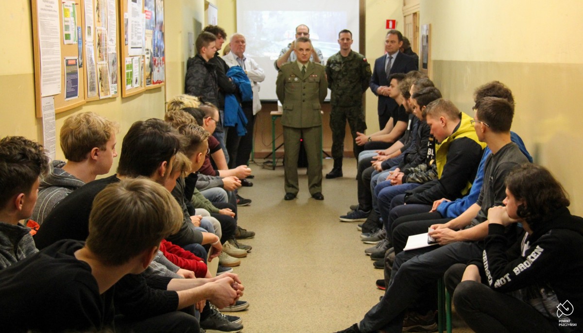 Wojskowy przemawiający do siedzących na ławkach mężczyzn.
