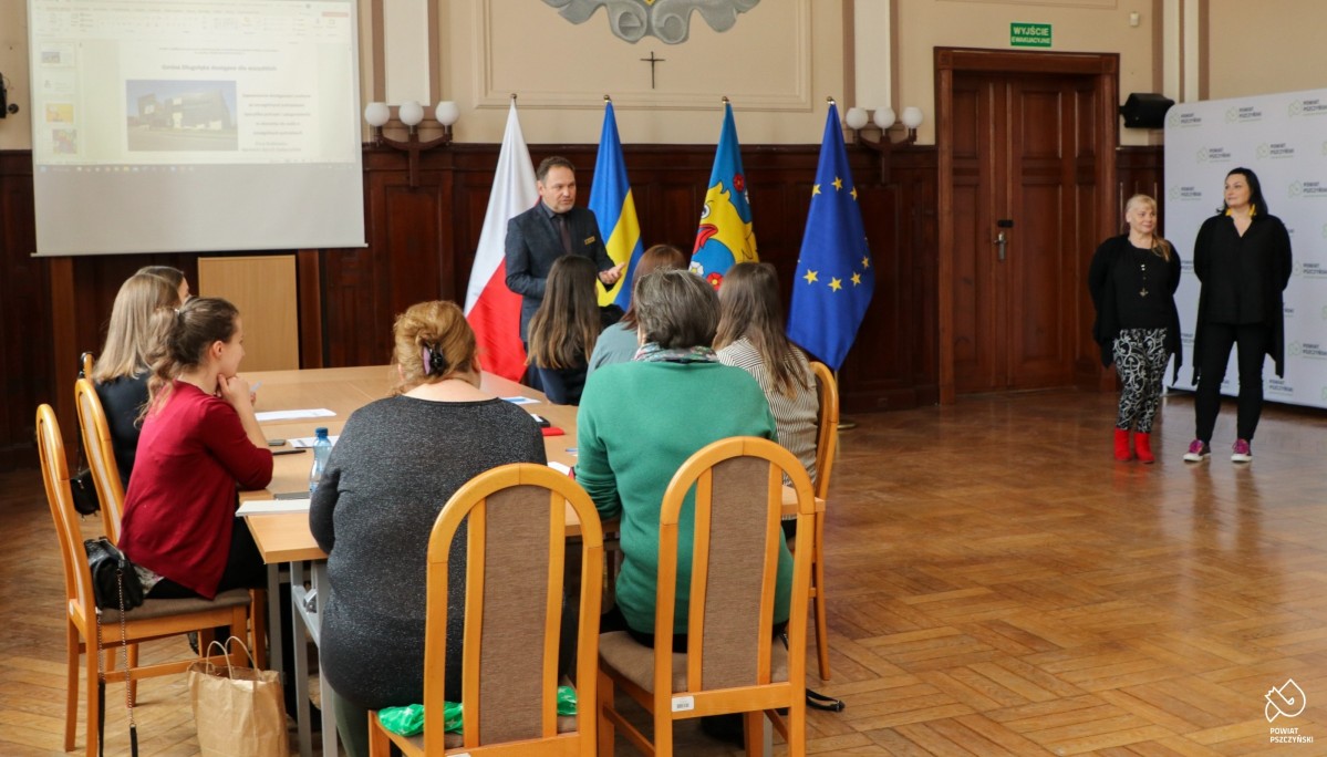 Grupa osób siedząca przy stole, przemawia wicestarosta. Obok kobiety prowadzące szkolenie.
