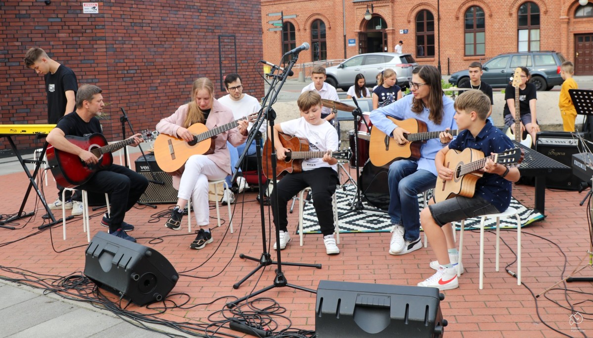 Uczniowie ogniska muzycznego siedzą na krzesłać na placu centrum przesiadkowego i grają na gitarach