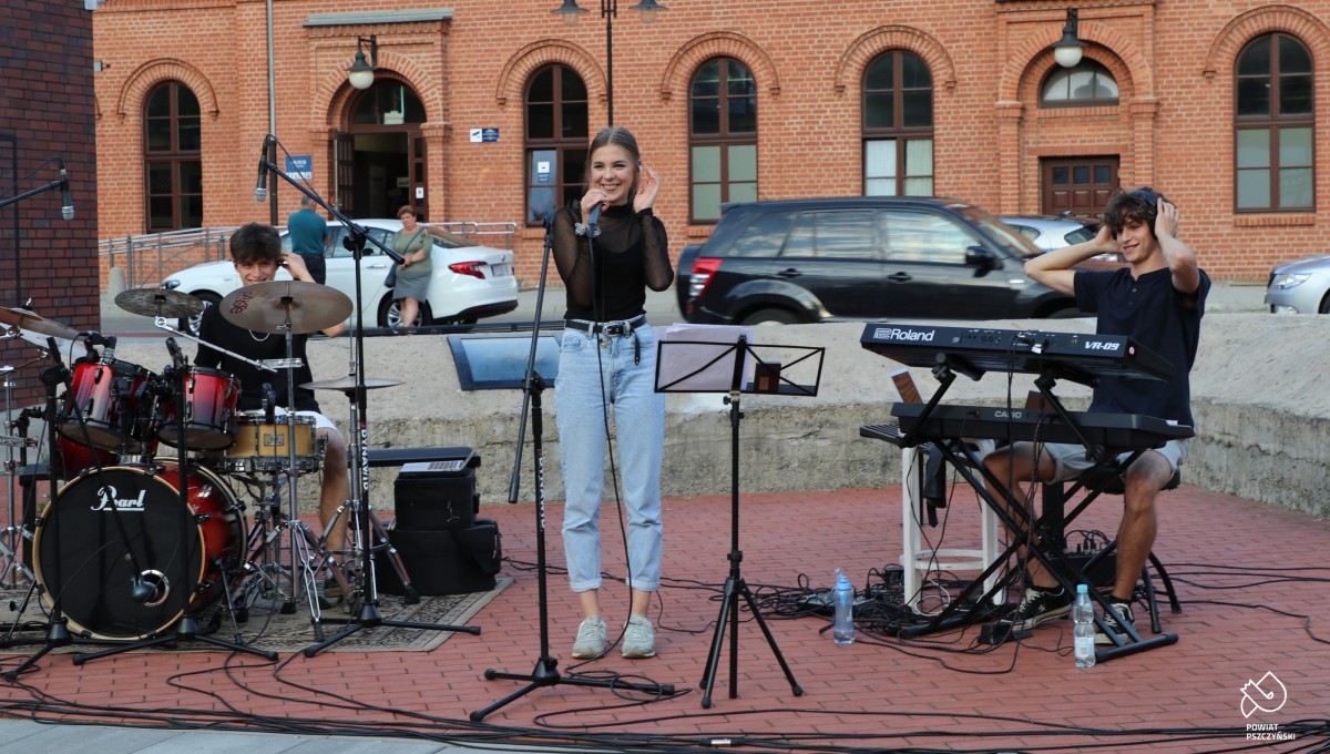 Na placu centrum przesiadkowego trwa koncert - śpiewa Zuzanna Piech, na klawiszach i perkusji grają członkowie zespołu B-L-P. W tle budynek dworca kolejowego.