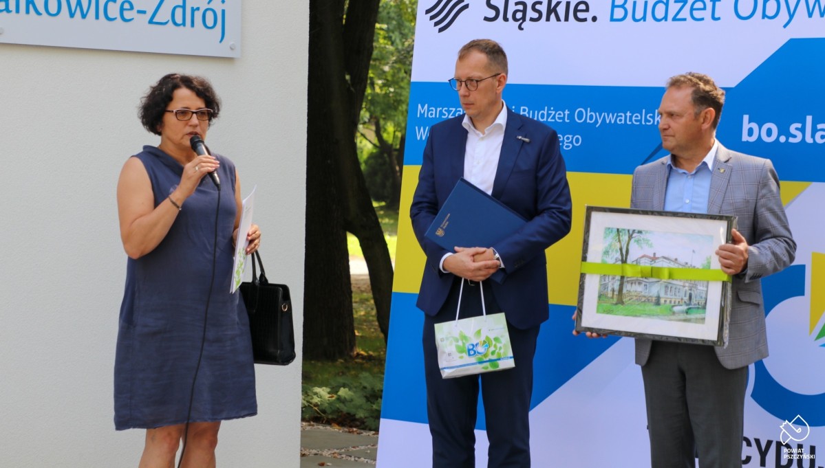 Starosta przemawia, obok stoi prezes uzdrowiska i wicestarosta, który trzyma grafikę.