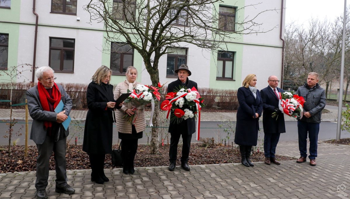 uczestnicy uroczystości stoją obok siebie, jedni trzymają w dłoniach wiązanki kwiatów.
