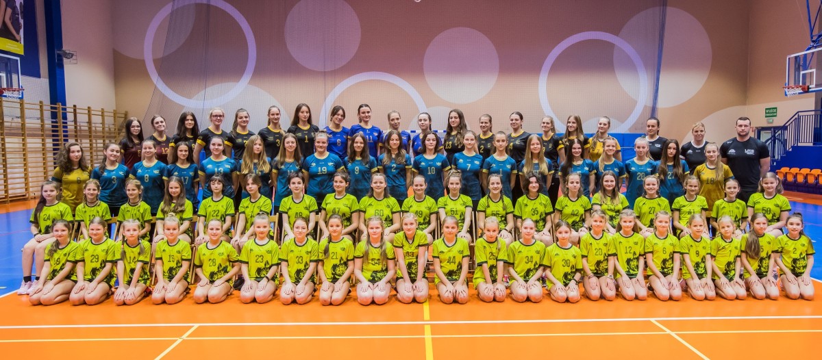 Zawodniczki szkółki siatkówki w pozowanym zdjęciu na hali sportowej. Są ubrane w żółte lub niebieskie stroje.