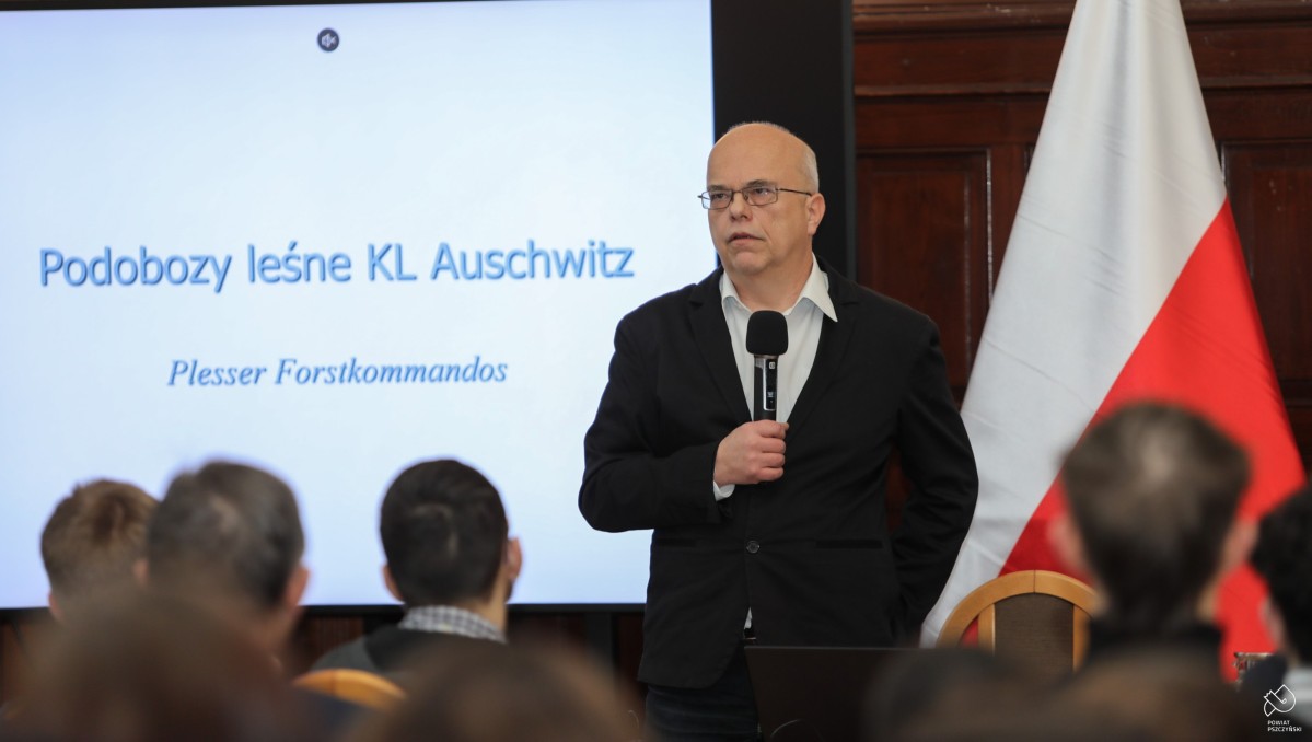 Mężczyzna prowadzący konferencję, przemawia, trzyma mikrofon, za nim wyświetlana jest prezentacja na ekranie telewizora.