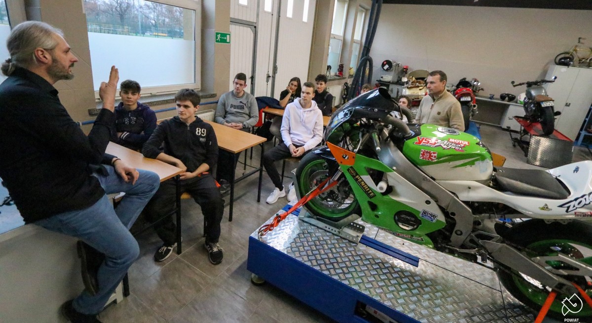 W pracowni motocyklowej uczniowie siedzą za stołami. Na pierwszym planie widoczny motocykl.