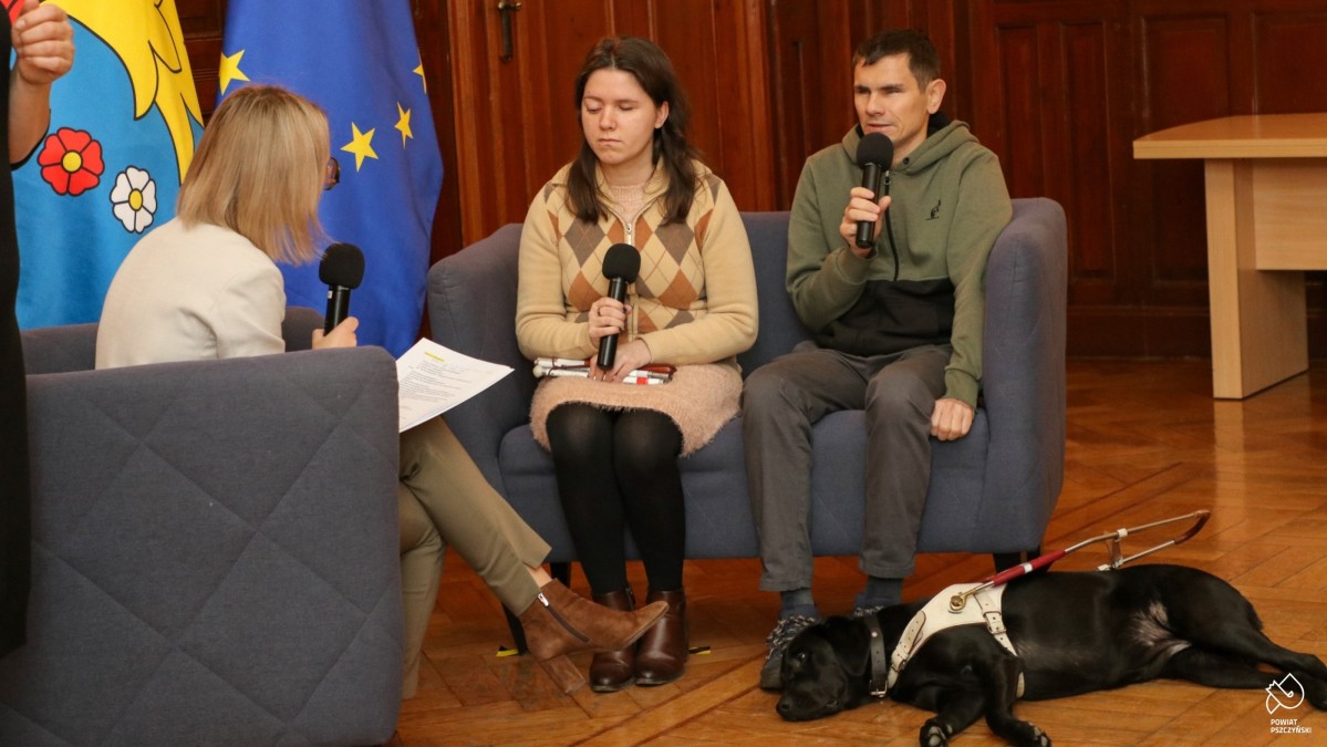 Rozmowa trzech osób, z których dwie to osoby niewidome. U stóp mężczyzny leży pies przewodnik.
