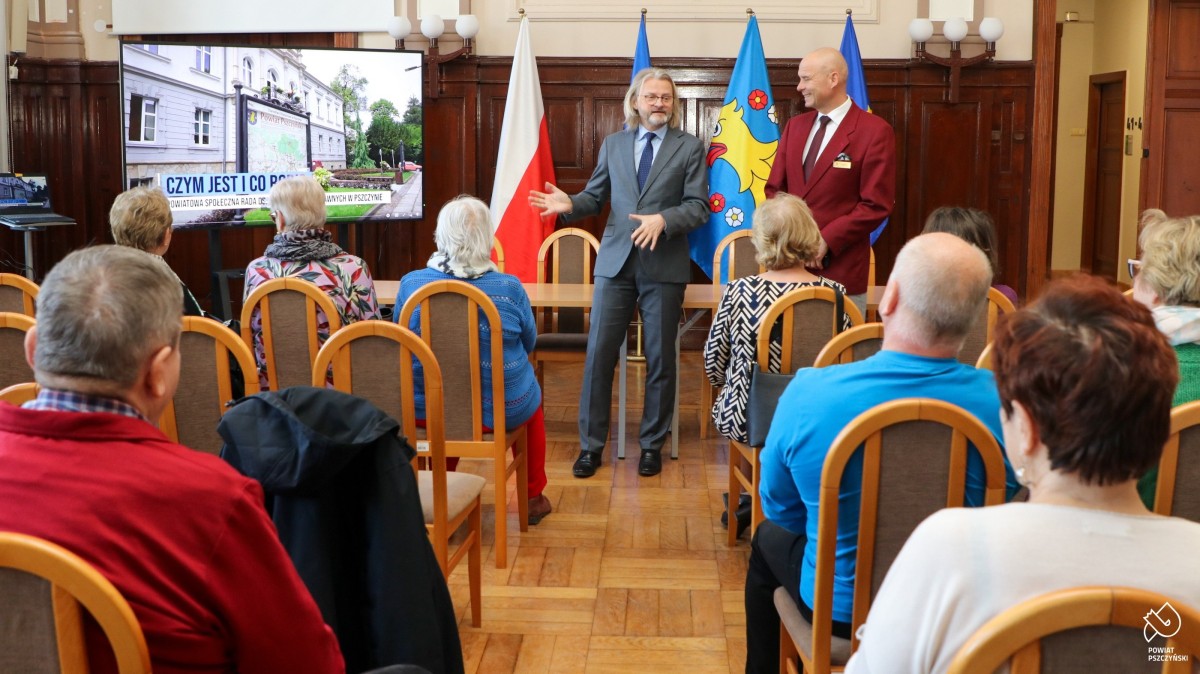 Przewodniczący Rady i sekretarz przemawiają do seniorów, którzy siedzą w sali sesyjnej.