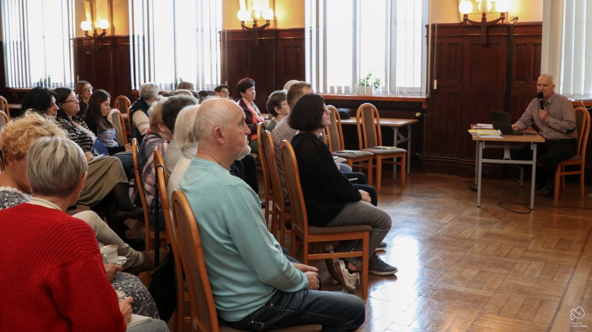 Uczestnicy spotkania siedzą na krzesłach w dużej sali, przed nimi siedzi prezenter.