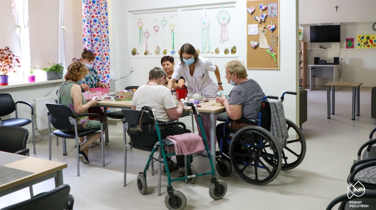 Jasne, duże pomieszczenie świetlicy, w którym przy stole siedzą seniorki oraz terapetki. Trwają zajęcia artystyczne.