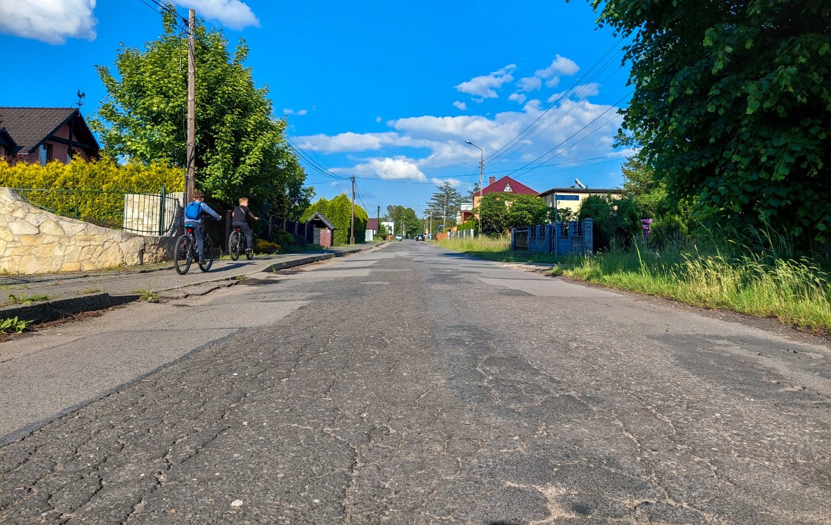 Fragment drogi, której nawierzchnia jest zniszczona. Chodnikiem poruszają się rowerzyści,