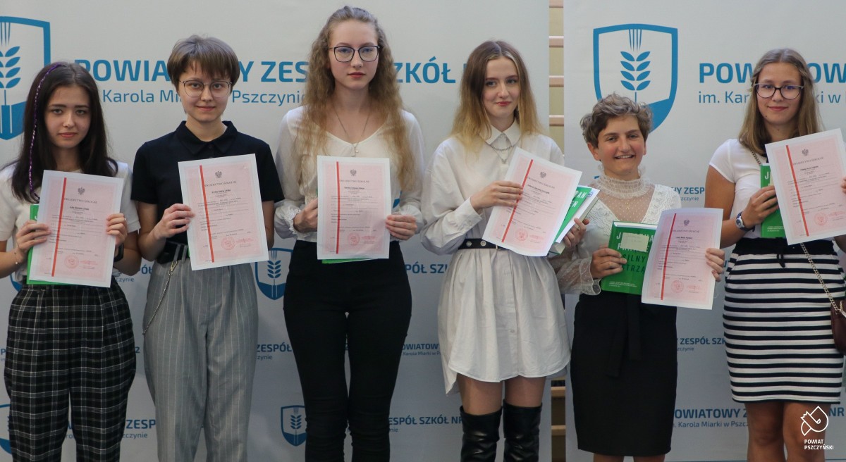 Uczennice stojące na tle ścianki z pzs nr 2 w pszczynie, w dłoniach trzymają świadectwa z czerwonym paskiem.