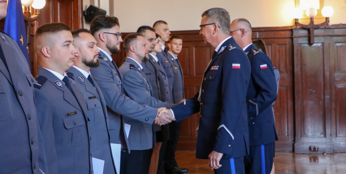 Kilku policjantów ubranych w policyjne mundury stoi obok siebie, a komendaci gratulują jednemu z nich wręczając białą teczkę z awansem.