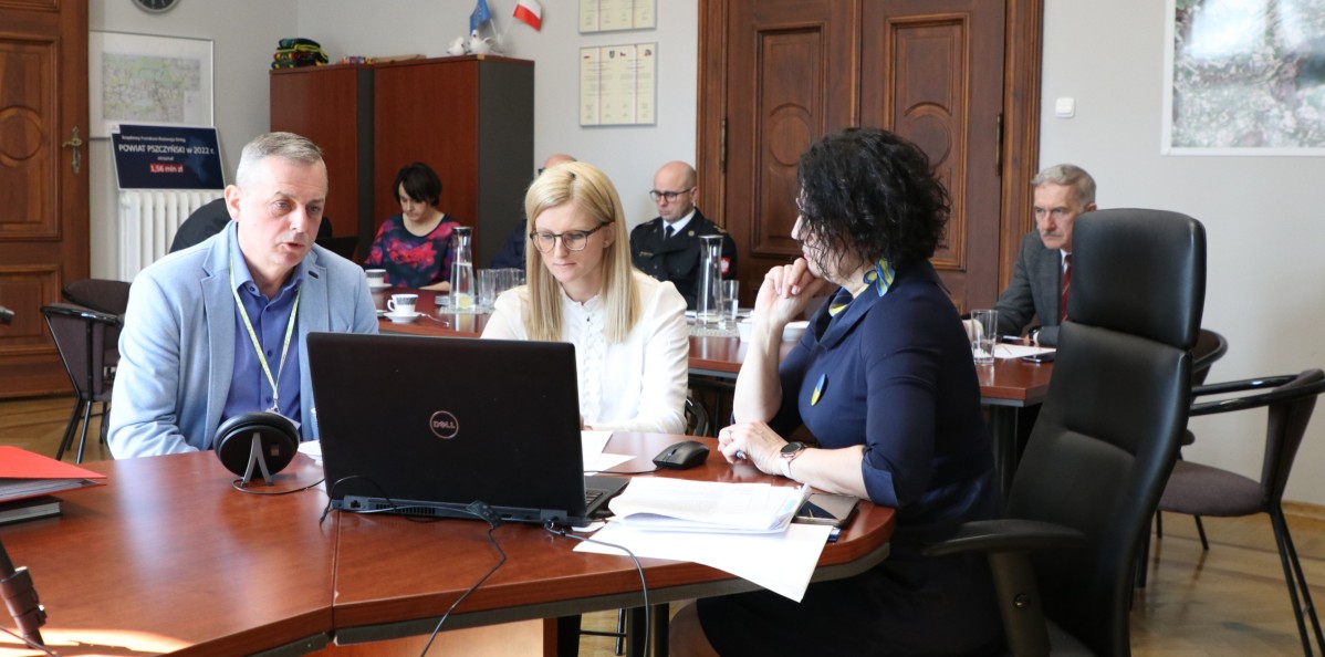 Za stołem przed komputerem siedzą trzy osoby. W tle znajdują się inne osoby