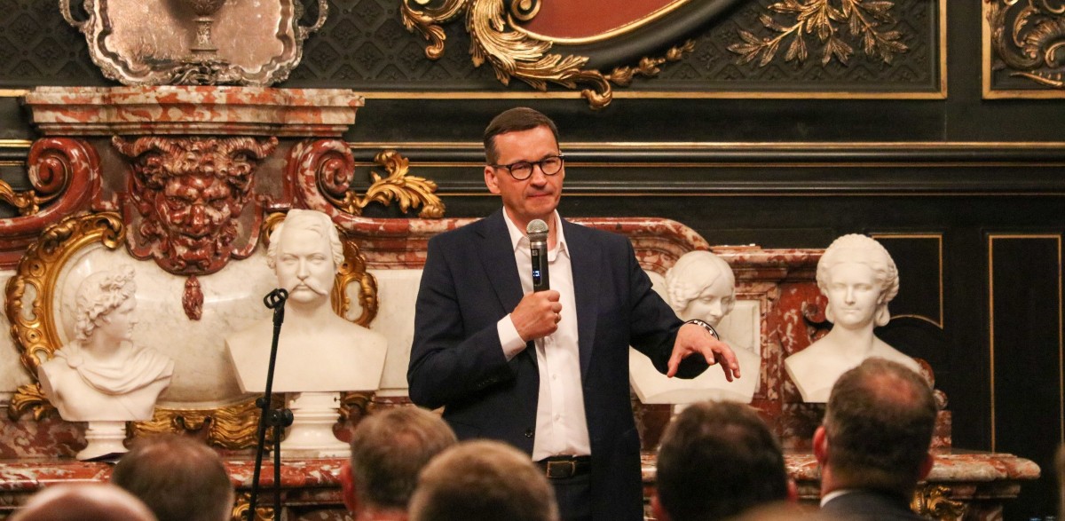 W sali lustrzanej pszczyńksiego zamku trwa spotkanie z premierem Mateuszem Morawieckim. Premier przemawia, trzymając mikrofon, na pierwszym planie głosy siedzących samorządowców.