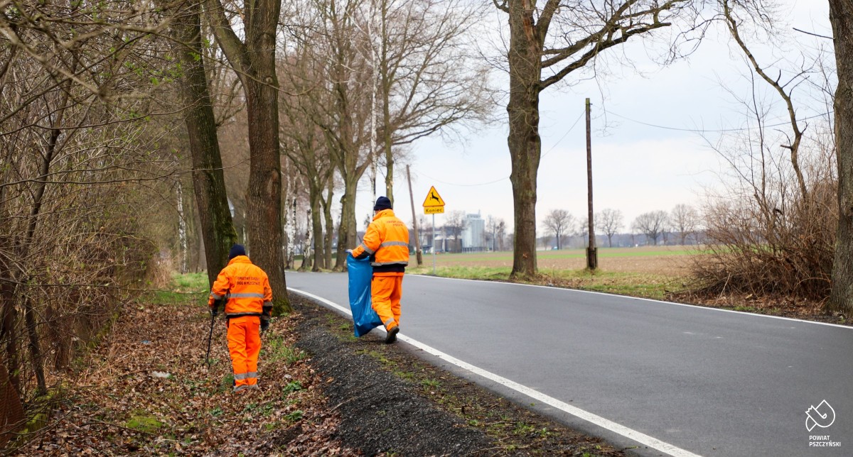 Mężczyźni ubrani w pomarańczowe stroje zbierają śmieci przy drodze.