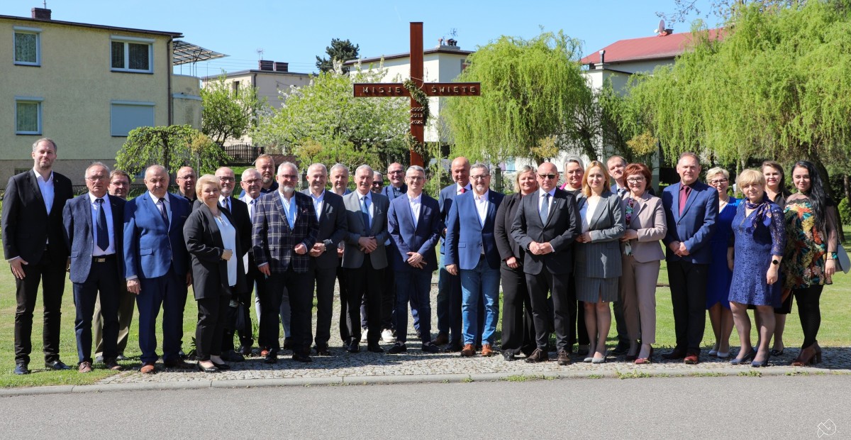 Grupa ok. 30 samorządowców stoi na terenie zielonym za nimi widoczny drewniany krzyż misyjny