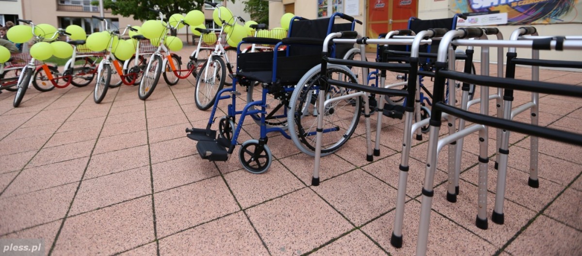 Sprzęt rehabilitacyjny ustawiony na zewnątrz. Widoczne wózki inwalidzkie, kule, chodziki oraz rowery trójkołowe.