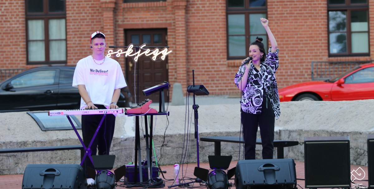 Koncert zepsołu sskjegg, kobieta z mikrofonem, mężczyzna gra na klawiszach, w tle dworzec kolejowy w Pszczynie