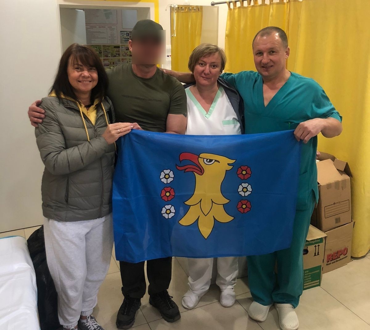 Cztery osoby - w tym medycy stoją w pomieszczeniu z flagą powiatu pszczyńskiego.