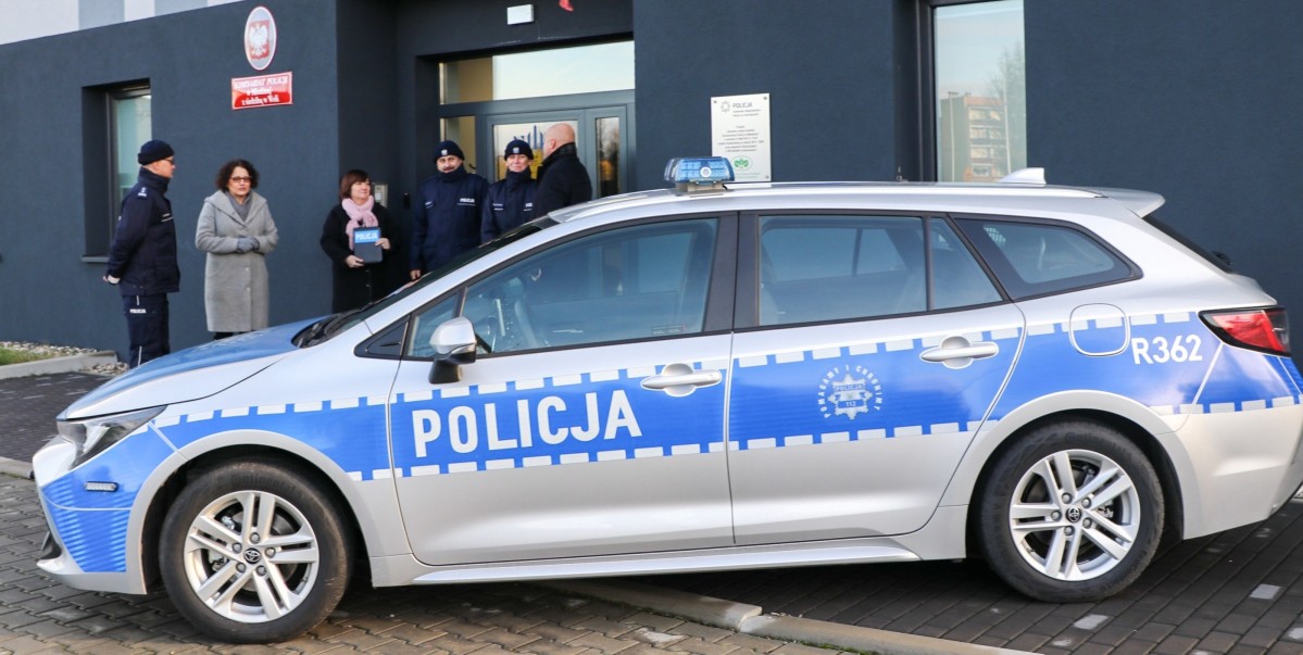 Przed budynkiem komisariatu stoi radiowóz policyjny, za nim stoi grupa ludzi.