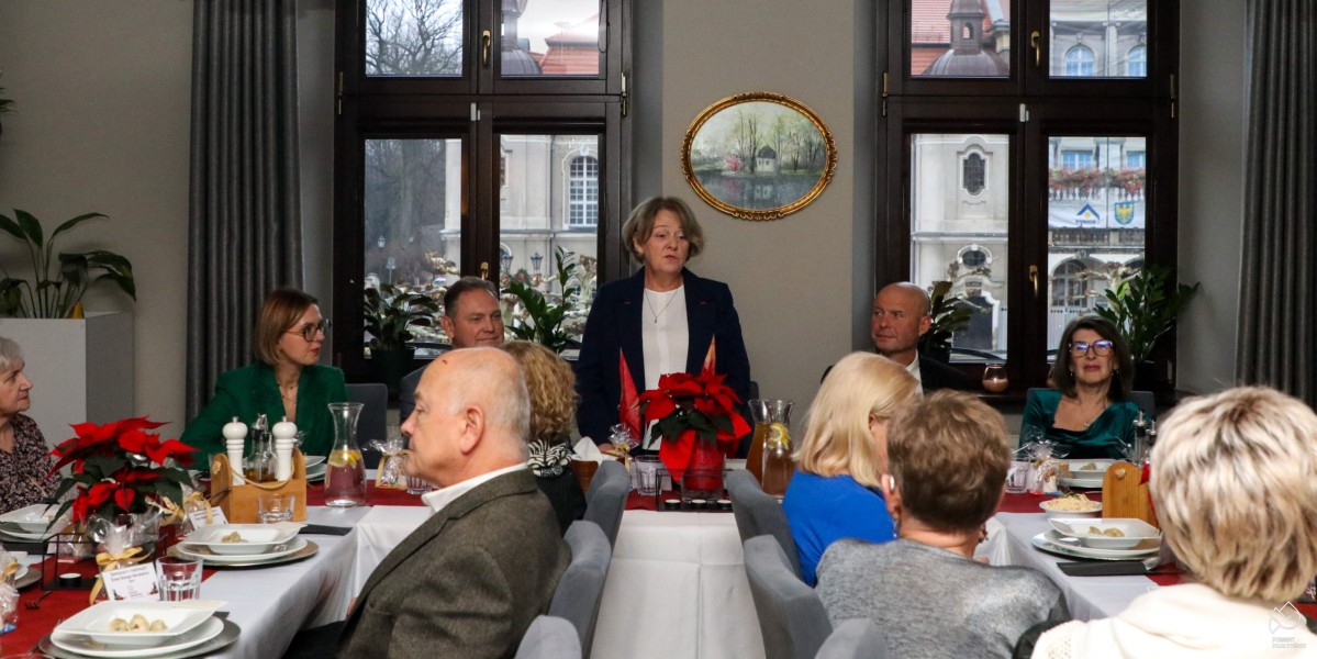 Przewodnicząca Forum stoi przy stole i przemawia, seniorzy siedzą przy wigilijnych stołach.