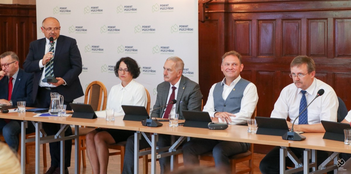 W sali sesyjnej powiatu podczas sesji za stołami siedzą członkowie Zarządu Powiatu, starosta stoi i trzyma mikrofon