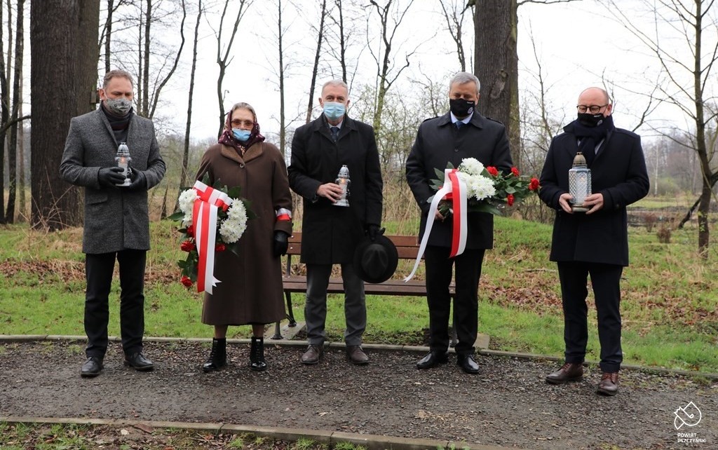 Czterech mężczyzn i jedna kobieta. Kobieta i jeden z mężczyzn w dłoniach trzymają biało-czerwone kwiaty. Pozostali mężczyźni trzymają zapalone znicze.