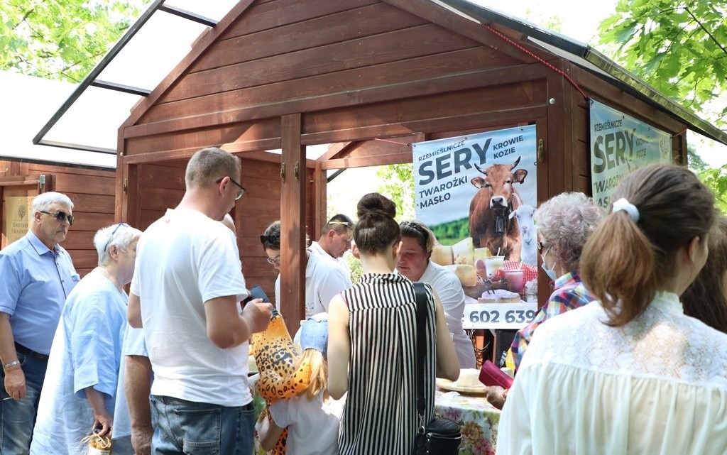 Drewniane stoisko, w którym promowane są naturalne sery podczas Festiwalu Lokalnych Produktów Ziemi Pszczyńskiej. Duże zainteresowanie ze strony przechodniów.