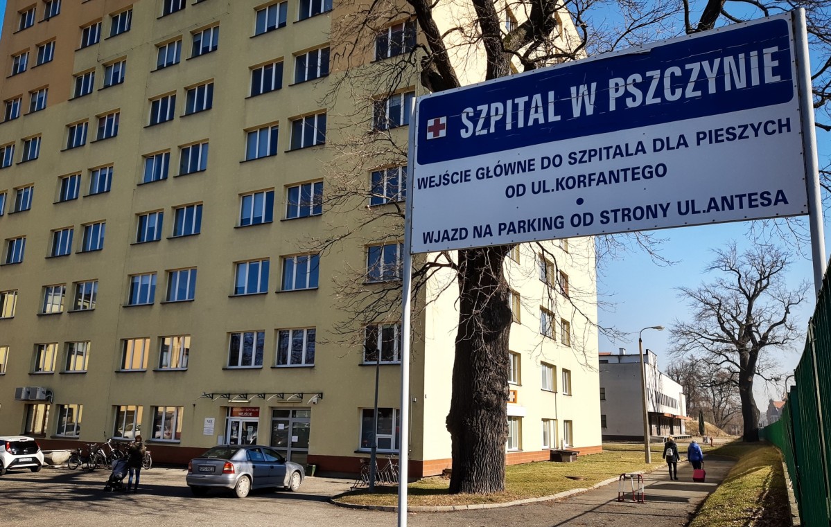 Budynek szpitala powiatowego w Pszczynie, wysoki, żółta elewacja. Obok tablica informacyjna o wejściu dla pieszych i wjeździe dla kierowców.
