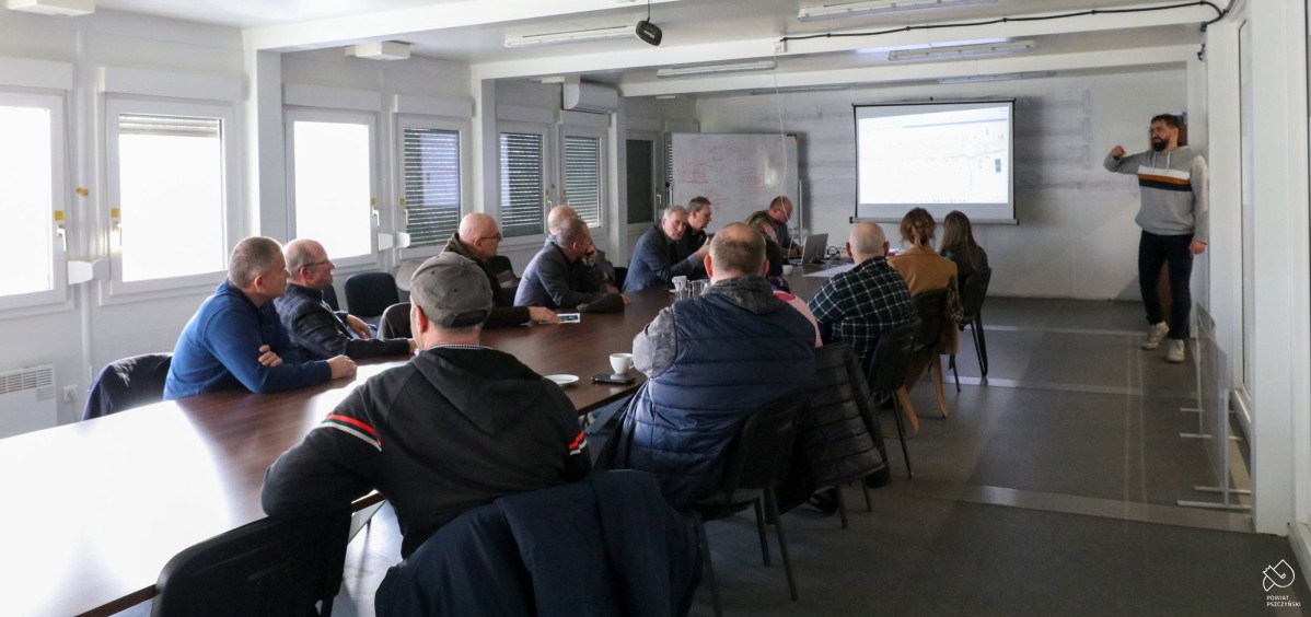 Podłużna duża sala w której przy długim stole siedzą uczetsnicy spotkania. Prowadzący spotkanie stoi z przodu przy ekranie, na którym wyświetlany jest projekt przebudowy przejazdu.