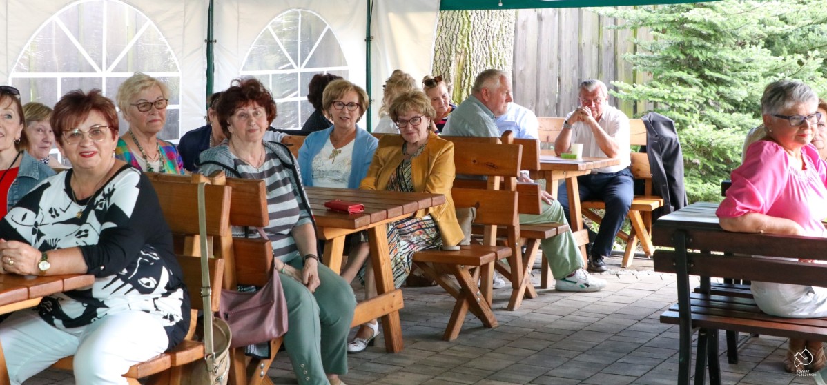 Grupa słuchaczu UTW siedzi za stołami pod namiotem w jankowickiej karczmie