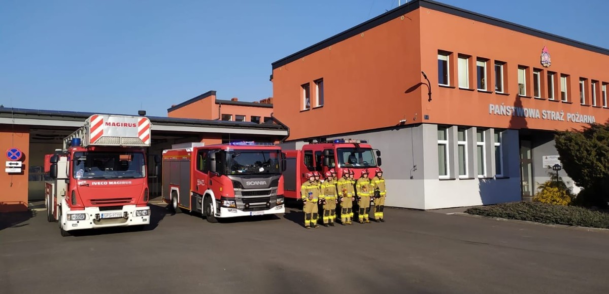 Przed budynkiem komendy straży pożarnej stoją trzy wozy strażackie oraz strażacy w żółtych ubranaich.