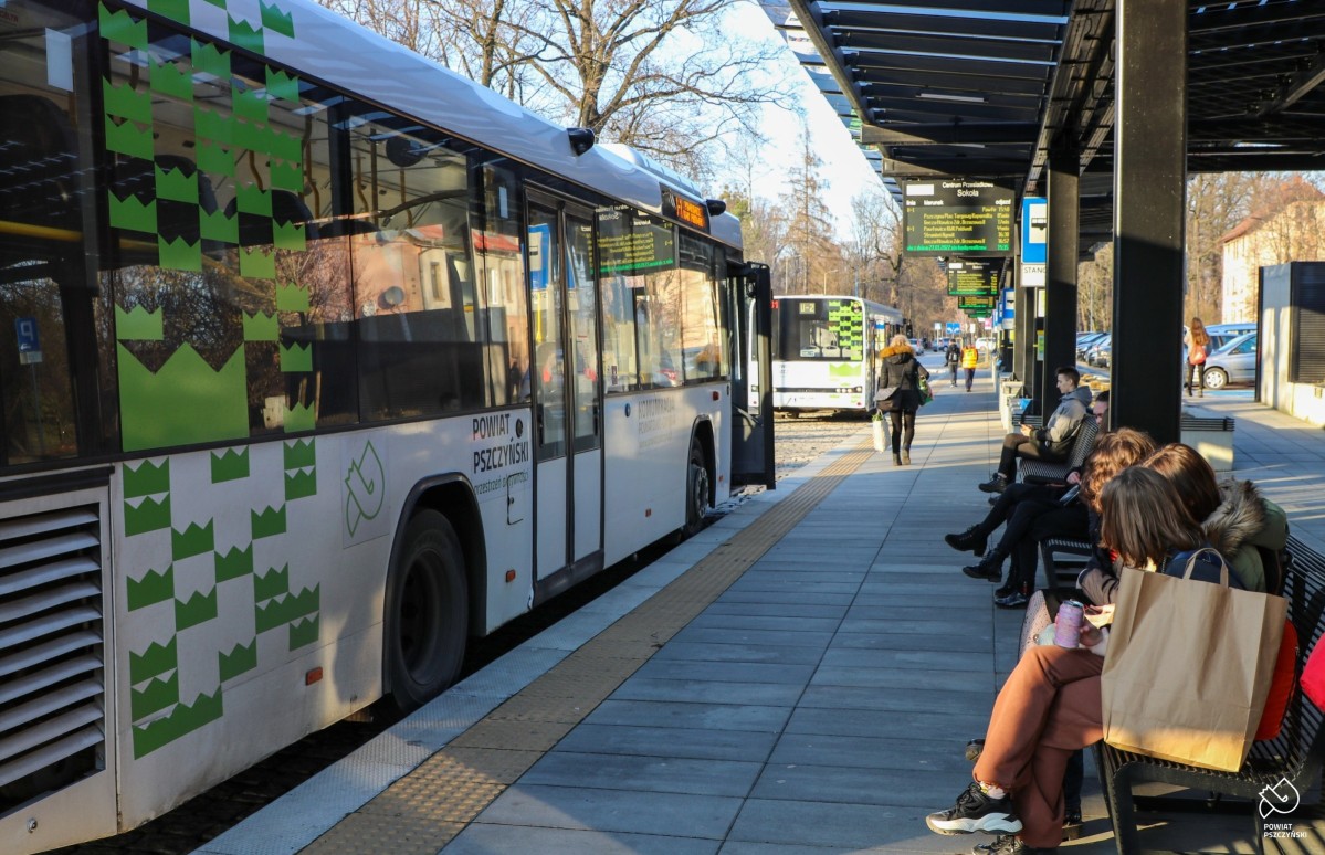 Biały autobus z zielonymi elementami graficznymi stoi przy peronie odjazdowym. Obok na ławeczkach siedzą pasażerowie.