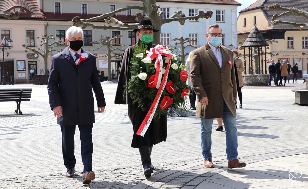 Wicestarosta, wiceprzewodniczący Rady Powiatu oraz skarbik powiatu pszczyńskiego niosą wiązankę kwiatów. W tle pszczyński rynek.