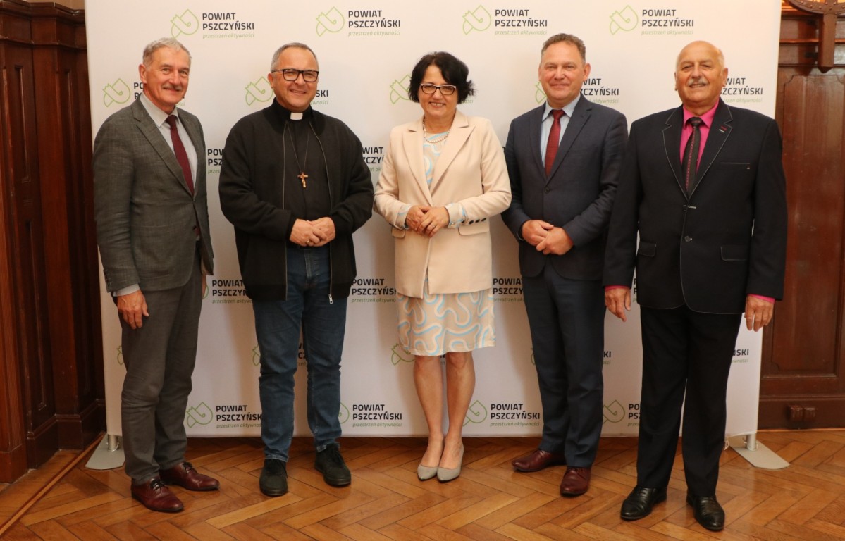 Włodarze powiatu przy ściance z logo powiatu z ks. bp Kałużą. Uśmiechają się.
