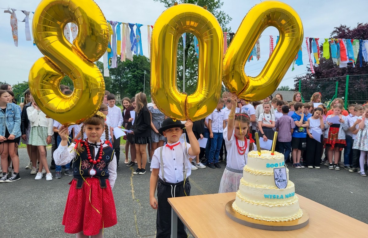 Dzieci ubrane w regionalne stroje trzymają balony z cyframi 8, 0, 0, przed nimi ustwiono piętrowy tort z herbem i napisem Wisła Mała.