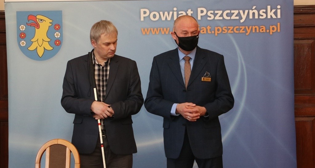 Dwaj mężczyźni stoją w dużej sali na tle niebieskiego baneru z napisem powiat pszczyński.
