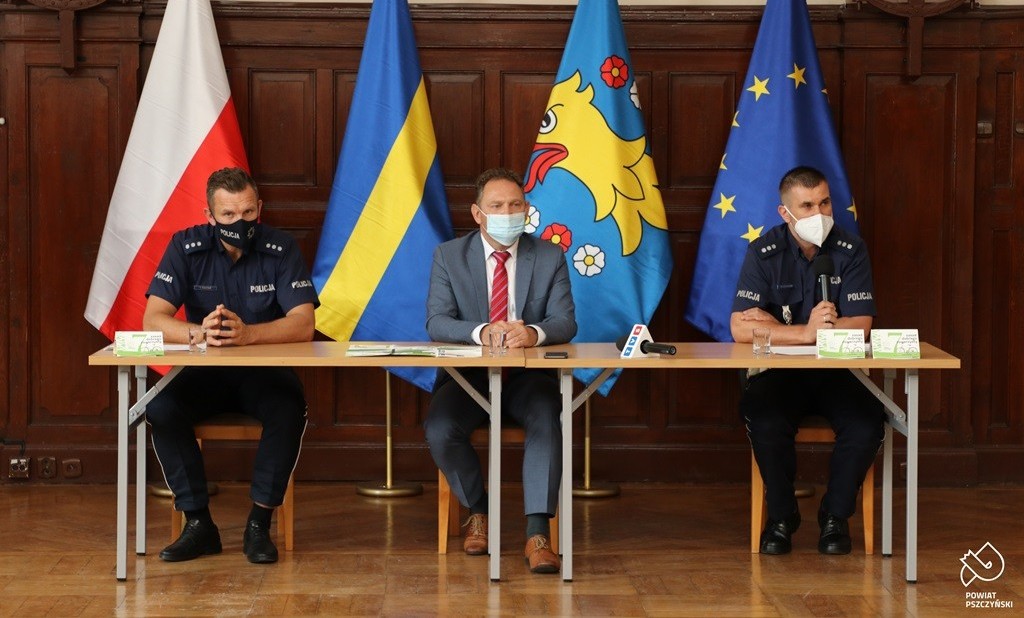 Policjant Komendy Wojewódzkiej Policji w Katowicach, wicestarosta pszczyński Damian Cieszewski i policjant Komendy Powiatowej Policji w Pszczynie. Siedzą za stołem. Za nimi stoją cztery flagi.
