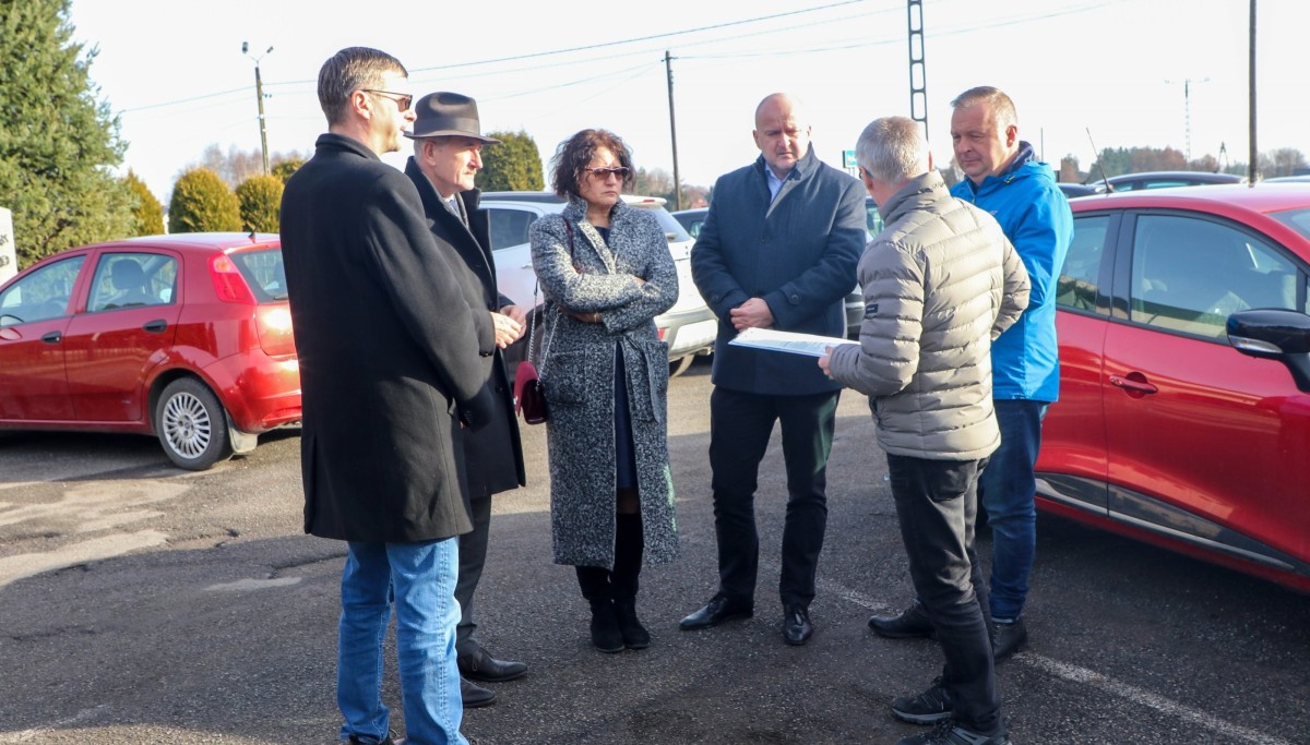 6 osób - przedstawiciele samorządów i wykonawcy stoją na parkingu obok ustawionych samochodów