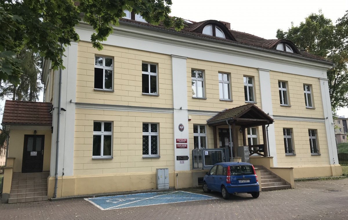 Grafika przedstawiająca budynek przy ul. Korfantego 6 w Pszczynie nazywany byłą Zarządcówką dwukondygnacyjny z żółto białą elewacją z dwoma wejściami przy głównym wejściu znajduje się podnośnik dla osób z niepełnosprawnościami