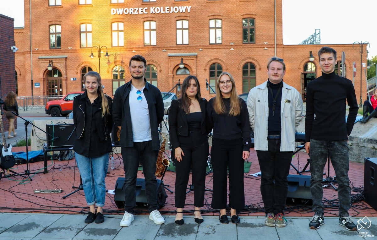 Członkowie zespołu Offbeat stojący w jednym rzędzie przed instrumentami na placu przed dworcem kolejowym.